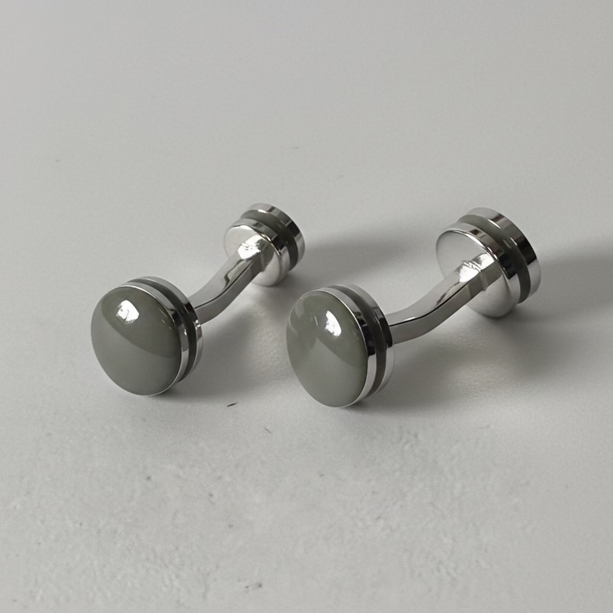 A pair of silver-toned cufflinks with round, polished dark gray to black front faces, photographed on a light gray textured surface.