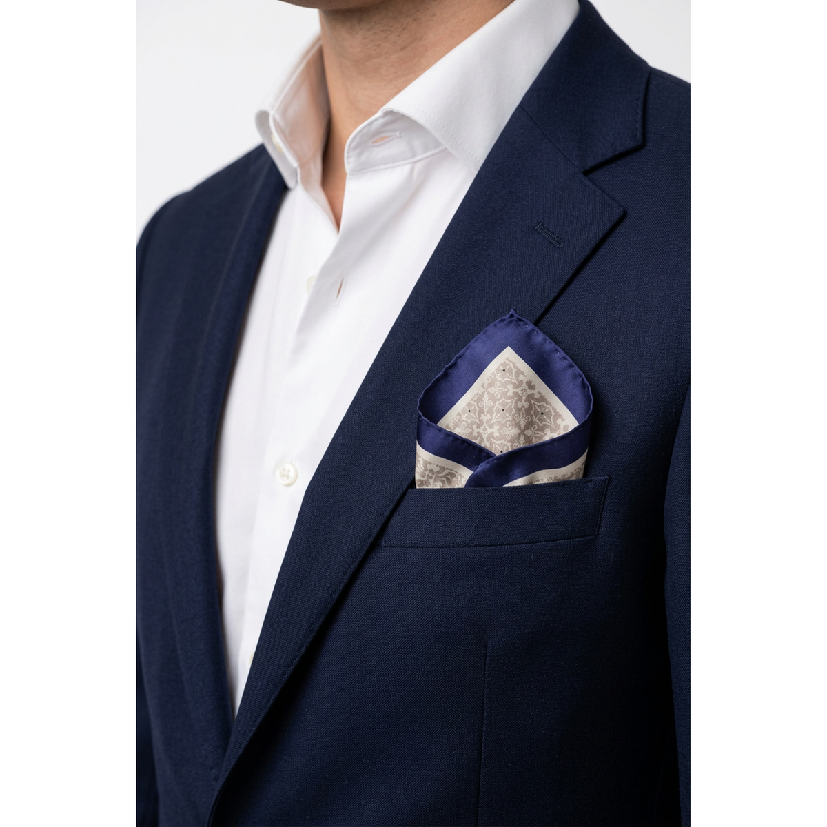 A close-up of a navy blue suit jacket worn by a model, paired with a white dress shirt and a patterned navy and beige pocket square in the breast pocket.