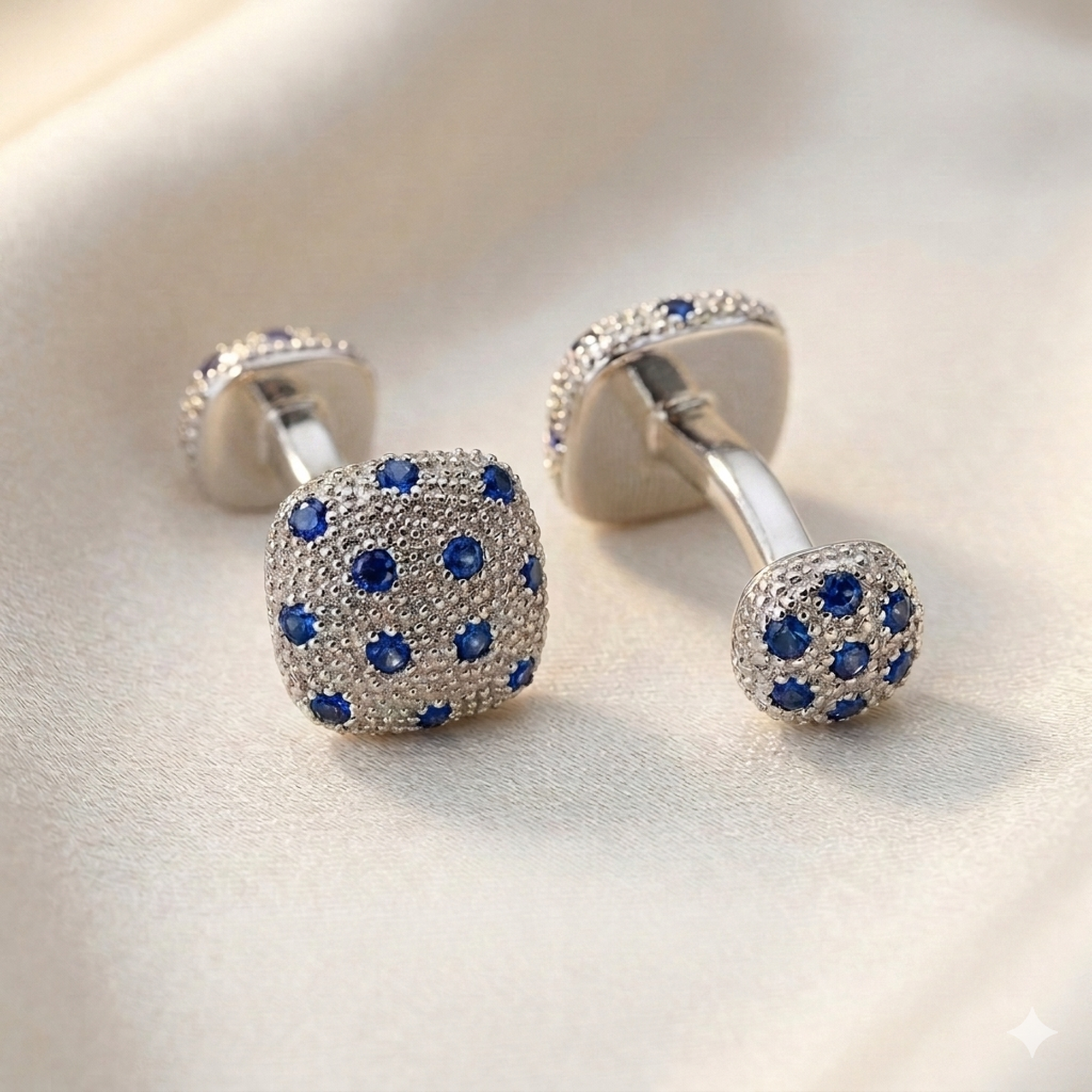 A pair of silver-toned cufflinks with square faces covered in small blue gemstones, photographed on a soft cream fabric background.